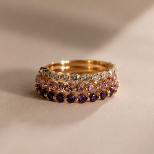 Puede incluir: Un conjunto de tres anillos de oro, cada uno con una hilera de piedras preciosas de corte redondo. El anillo superior tiene piedras blancas claras, el central piedras rosa claro y el inferior piedras moradas oscuras, montadas en una banda de metal.