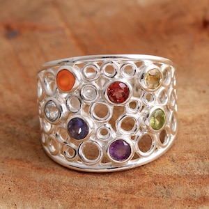 Può includere: Un anello d'argento con un design traforato unico, caratterizzato da una varietà di pietre preziose colorate. L'anello presenta una miscela di pietre rotonde nelle tonalità arancione, rosso, giallo, verde, viola e blu, incastonate in una fascia d'argento strutturata.