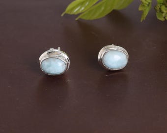 May include: A pair of oval-shaped stud earrings. Each earring features a light blue gemstone set in a silver-colored metal. The earrings are displayed on a brown surface, with green leaves in the background.