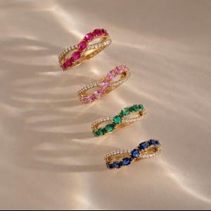 May include: Four gold rings arranged in a diagonal line, each featuring an infinity-shaped band. The rings are set with small clear stones and larger oval-cut gemstones in red, light pink, green, and blue, creating a vibrant and colorful collection.