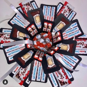 May include: A black and white explosion box filled with Kinder Chocolate bars and sweets. The box is decorated with a white ribbon and the words "Kinder Bueno" are visible on the chocolate bars.