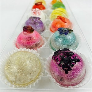 May include: A dozen colorful, dome-shaped confections in a clear plastic tray. Each treat is a different color, including pink, blue, yellow, and purple, and is decorated with edible flowers or other toppings. The treats sit in white paper cups.