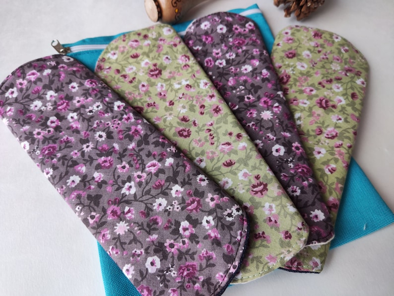 May include: Four reusable cloth pads with floral patterns in shades of green, purple, and white. The pads are arranged in a fan shape and are sitting on a blue fabric pouch.