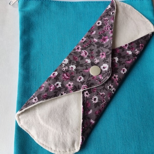 May include: A reusable cloth menstrual pad with a floral print on a turquoise background. The pad is folded in half and has a white button on the front.