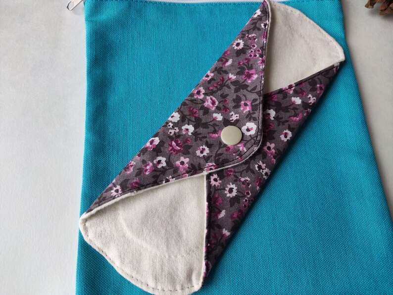May include: A reusable cloth menstrual pad with a floral print on a turquoise background. The pad is folded in half and has a white button on the front.