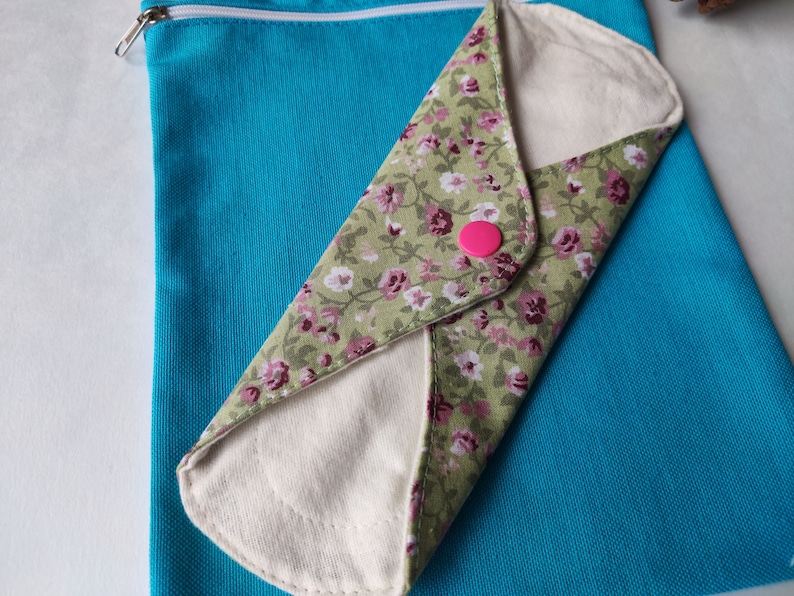 May include: A reusable cloth menstrual pad with a floral print, folded and placed inside a turquoise zippered pouch.
