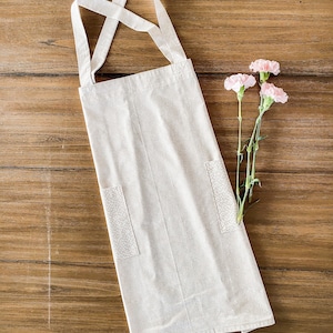 Puede incluir: Un delantal de algodón blanco con un diseño de correa cruzada y dos bolsillos. El delantal tiene un sutil patrón de lunares blancos.