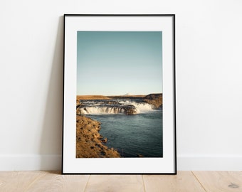 May include: A framed photograph of a waterfall cascading over rocks into a turquoise pool. The sky is a pale blue, and a mountain peak is visible in the distance.