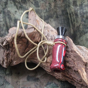 May include: A red and black wooden duck call with a black metal mouthpiece and a tan lanyard. The call is resting on a piece of driftwood.