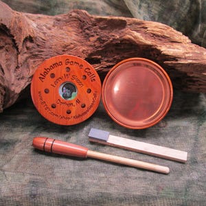 May include: A wooden game call set with a copper-colored lid and a striker. The call is round with the text "Alabama Game Calls" and "Larry W. Skeen" engraved on it. The set includes a wooden striker and a small, rectangular piece of wood with a blue top.