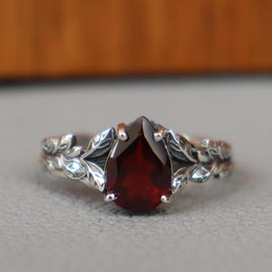 May include: A silver ring with a pear-shaped, deep red gemstone. The ring features a leaf design on the band and around the gemstone. The gemstone is the focal point, set in a detailed, nature-inspired setting.