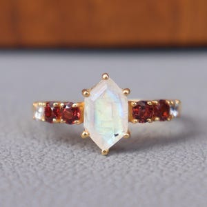 Może przedstawiać: Złoty pierścionek z centralnym, iryzującym, sześciokątnym kamieniem szlachetnym. Opaska ozdobiona jest naprzemiennie czerwonymi i przezroczystymi kamieniami. Pierścionek umieszczony jest na neutralnej szarej powierzchni, z drewnianym tłem.