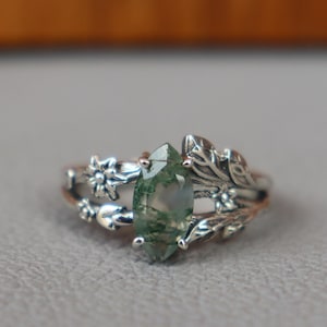 Puede incluir: Un anillo de plata con una gema verde en forma de marquesa, adornado con detalles florales y de hojas. La banda del anillo presenta pequeñas flores y hojas, creando un diseño inspirado en la naturaleza. La gema verde es el punto focal.