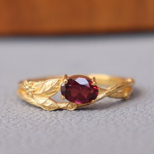 May include: A gold ring featuring a deep red oval gemstone. The ring's band is adorned with leaf-like details, creating a nature-inspired design. The gemstone is the focal point, set in a delicate, ornate setting.
