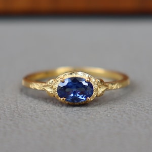 Könnte beinhalten: Ein goldener Ring mit einem ovalen, tiefblauen Edelstein. Der Ring hat ein zartes, strukturiertes Band und eine verzierte Detaillierung um die Edelsteinfassung. Der Ring ist vor einem neutralen grauen Hintergrund platziert.