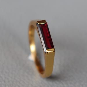 May include: A gold ring with a rectangular, deep red gemstone. The ring has a simple, elegant design with a polished finish. The gemstone is set flush within the gold band, creating a sleek, modern look. The ring is shown against a neutral grey background.