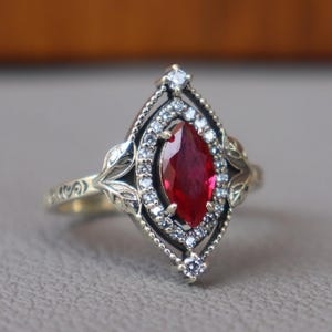 May include: A gold-toned ring featuring a marquise-cut, deep red gemstone at its center, surrounded by smaller white gemstones. The ring has an ornate, filigree design with leaf-like details on the band and around the central stone.