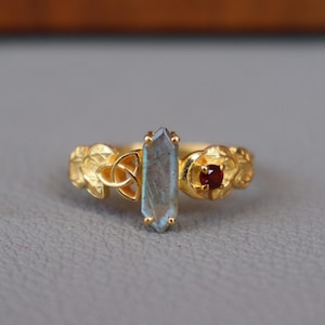 May include: A gold-colored ring featuring a light blue crystal and a small red gemstone. The ring has a detailed floral design with a Celtic knot. The ring is set on a gray surface.
