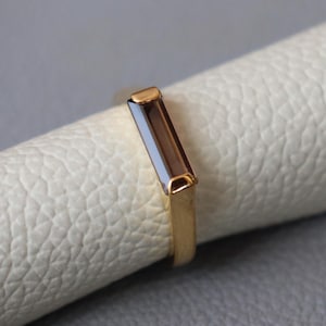 May include: A gold ring featuring a single rectangular baguette-cut brown gemstone set horizontally. The band is smooth and polished, showcasing a clean metal finish that complements the deep, translucent brown hue of the central stone.