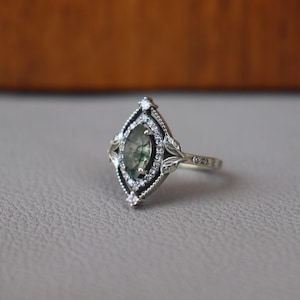May include: A silver ring with an oval, green-hued stone at the center, surrounded by small, clear stones. The ring has a detailed, ornate design with leaf accents and a textured band. The setting is a marquise shape.