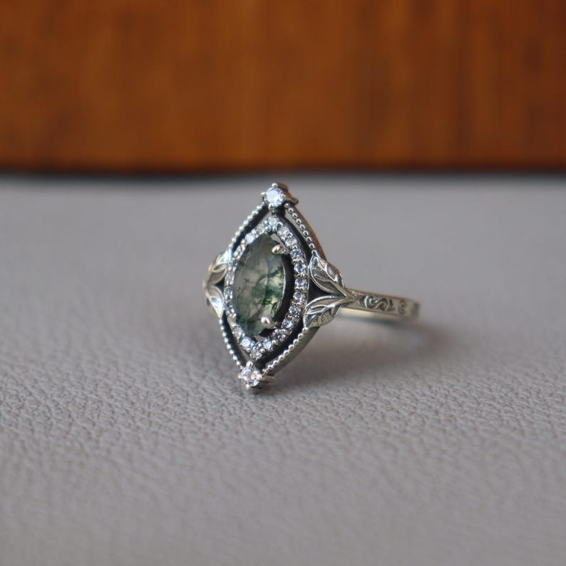 May include: A silver ring with an oval, green-hued stone at the center, surrounded by small, clear stones. The ring has a detailed, ornate design with leaf accents and a textured band. The setting is a marquise shape.