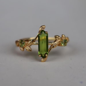 May include: A gold ring featuring a central vertical baguette-cut green gemstone flanked by two smaller round green gemstones. The band is designed with textured gold leaves and vine-like metalwork extending from the central stone setting.