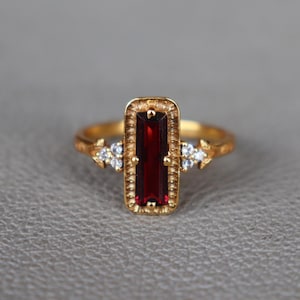 May include: A gold ring featuring a central rectangular baguette-cut deep red garnet stone set within a textured frame. The ring band is accented on each side by a cluster of three small, clear, round-cut white stones set in gold metal.
