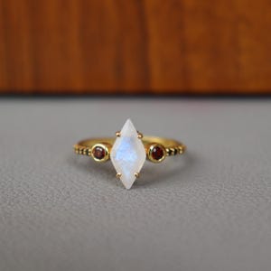 May include: A gold ring featuring a central, kite-shaped, iridescent moonstone. Two small, round, red gemstones flank the moonstone. The band has a textured design. The ring is set against a gray surface.