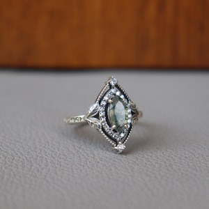 May include: A silver ring with an oval, green gemstone at the center, surrounded by small white stones. The ring has a detailed, leaf-like design around the gemstone and on the band. The ring is set on a gray surface.