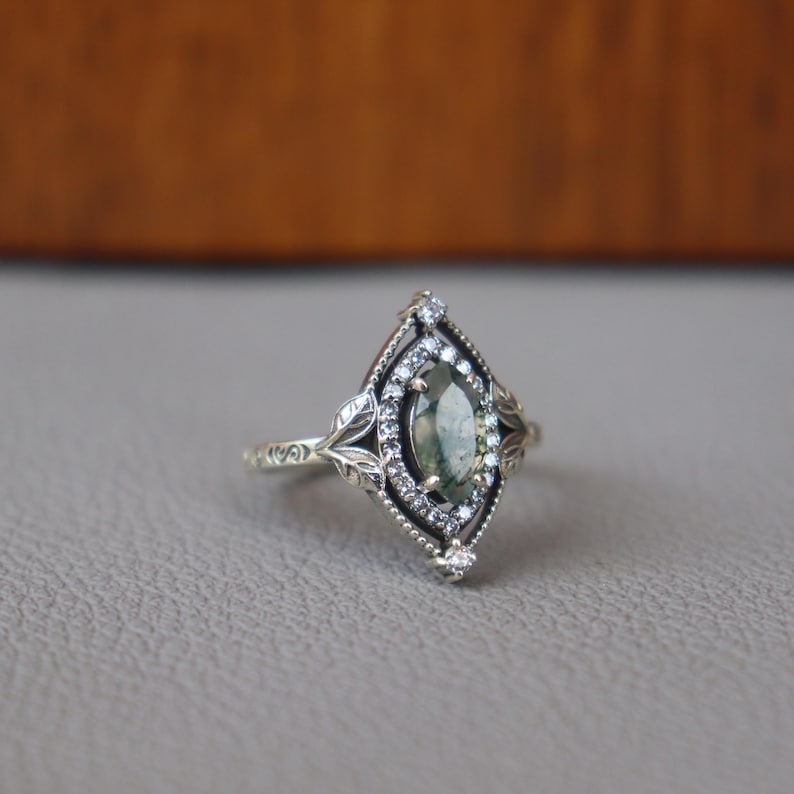 May include: A silver ring with an oval, green gemstone at the center, surrounded by small white stones. The ring has a detailed, leaf-like design around the gemstone and on the band. The ring is set on a gray surface.