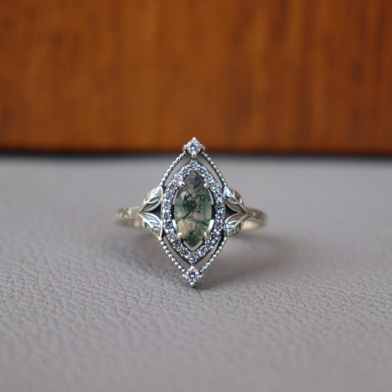 May include: A silver ring with an oval, moss agate stone at the center. The stone is surrounded by small, clear gemstones and a decorative filigree design. Leaf-like details adorn the sides of the ring, adding to its vintage aesthetic.