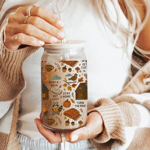 May include: A clear glass tumbler with a bamboo lid and straw, decorated with fall-themed illustrations and text. The design includes pumpkins, leaves, and the phrases "Read a Book" and "Stay Home".