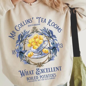 Może przedstawiać: Beżowa bluza z niebiesko-białym nadrukiem graficznym przedstawiającym filiżankę herbaty z cytrynami i tekstem "Mr Collins' Tea Rooms Hertfordshire 1813 What Excellent Boiled Potatoes A Delightful Spot for Tea & Company".