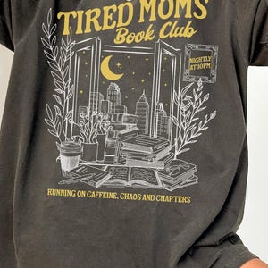 Op de afbeelding: Donkergrijze oversized t-shirt met een grafisch ontwerp. Het ontwerp toont de tekst "Tired Moms Book Club" boven een illustratie van een stadsgezicht gezien door een raam, met boeken en planten. De tekst "Running on Caffeine, Chaos and Chapters" staat onderaan.