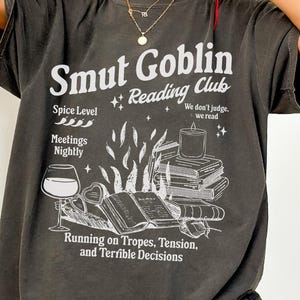 May include: A dark grey t-shirt with a white graphic print that says "Smut Goblin Reading Club". The graphic includes a book, a candle, a glass of wine, and the text "Spice Level", "Meetings Nightly", "We don't judge, we read", and "Running on Tropes, Tension, and Terrible Decisions".
