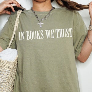 May include: A sage green oversized t-shirt with the text "In Books We Trust" printed in white on the front. The t-shirt is being worn by a person holding a woven straw bag.