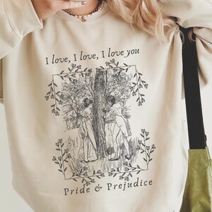 Puede incluir: Una sudadera beige con una ilustración en blanco y negro de dos personas de pie bajo un árbol. El texto "I love, I love, I love you" y "Pride & Prejudice" están impresos en la sudadera.