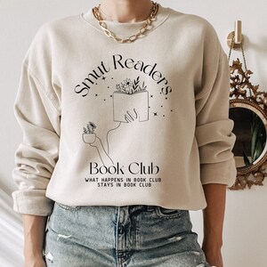 May include: A beige sweatshirt with the text "Smut Readers Book Club" and a graphic of a hand holding a book with flowers. The text below the graphic reads "What happens in book club stays in book club."