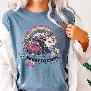 May include: A blue t-shirt featuring a possum, roses, and a rainbow design. The text "BE GAY DO CRIME" is displayed on a banner. The shirt is paired with denim shorts.