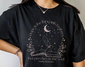 May include: Black t-shirt featuring a quote by Emily Dickinson: "Hope is the thing with feathers that perches in the soul." The design includes a crescent moon, stars, a feather, and an open book. The text and graphics are in a neutral color.