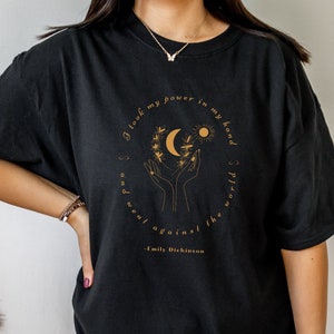 May include: Black oversized t-shirt with a gold graphic of hands holding a crescent moon and sun with a quote from Emily Dickinson: "I took my power in my hand and went against the world."