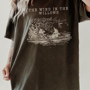 Puede incluir: Camiseta verde oliva oscuro con el texto "THE WIND IN THE WILLOWS" y una ilustración de una escena de río. La imagen muestra personajes y una libélula. La camiseta tiene mangas cortas y un corte holgado.