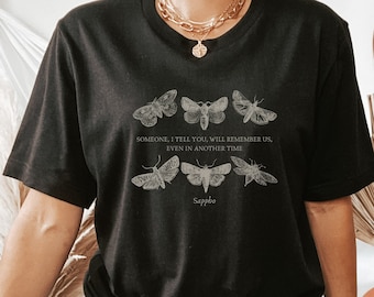 May include: Black t-shirt featuring a design of six moths and the text "SOMEONE, I TELL YOU, WILL REMEMBER US, EVEN IN ANOTHER TIME Sappho." The shirt is a dark color and has a classic crew neck.