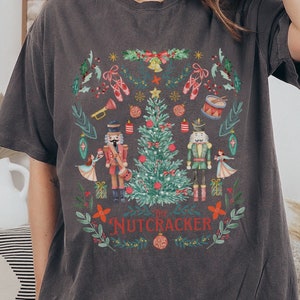 May include: Dark gray t-shirt featuring a colorful Christmas-themed graphic. The design includes a Christmas tree, nutcrackers, ballet shoes, and the text "The Nutcracker" in red. The shirt is a comfortable, casual style.