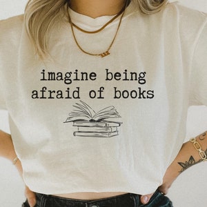 Op de afbeelding: Een wit T-shirt met de tekst "imagine being afraid of books" in zwart gedrukt. Eronder is een stapel boeken met een open boek bovenop afgebeeld.