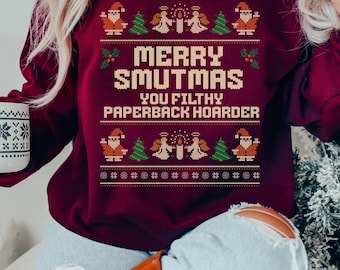 May include: Burgundy sweatshirt with a festive design. The text "MERRY SMUTMAS YOU FILTHY PAPERBACK HOARDER" is displayed in a pixelated font. The design includes Santa Claus, angels, and Christmas trees. A white mug with snowflake designs is held in the left hand.