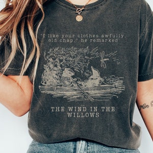 May include: A dark gray t-shirt with a vintage-style illustration of two characters in a boat. The text "I like your clothes awfully. old chap," he remarked" and "THE WIND IN THE WILLOWS" is printed on the shirt.