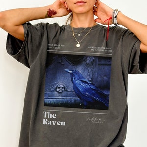 May include: A dark gray oversized t-shirt with a graphic print of a raven on a tombstone. The text "The Raven" is printed below the image. The text "Edgar Allan Poe 1809 - 1849" and "American writer, poet, and literary critic" is printed above the image.