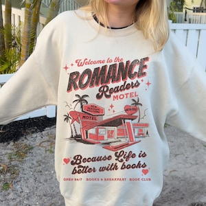 May include: A beige sweatshirt with a graphic print of a motel called "Romance Readers Motel". The graphic features a retro motel with palm trees and the text "Welcome to the Romance Readers Motel" and "Because Life is better with books".