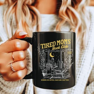 Może przedstawiać: Czarny ceramiczny kubek z napisem "Tired Moms Book Club" oraz grafiką przedstawiającą panoramę miasta, księżyc i książki. Kubek ma również napis "Running on coffee, chaos and chapters".